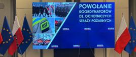 Monitor z banerem na którym widnieje w prawym górnym rogu napis POWOŁANIE KOORDYNATORÓW DS. OCHOTNICZYCH STRAŻY POŻARNYCH, po lewej stronie zdjęcia strażaków i samochodów pożarniczych