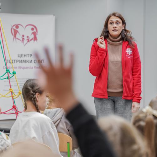 Kobieta w czerwonym polarze z napisem Caritas prowadzi zajęcia dla grupki dzieci odwróconych tyłem na zdjęciu, widać 1 uniesioną rękę