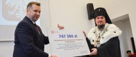 Minister Czarnek i mężczyzna w czarnym ceremonialnym stroju trzymają wielki symboliczny czek z napisem 747 380 zł.