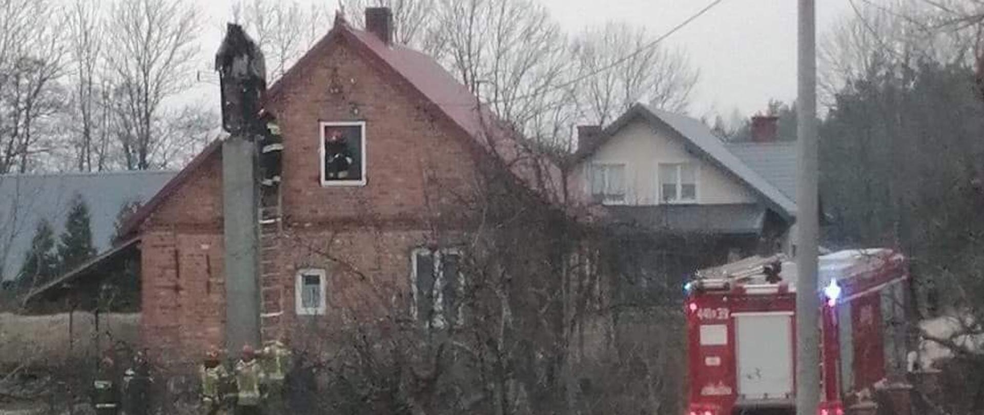 Na zdjęciu widać dom w którym doszło do pożaru w przewodzie kominowym. W oknie domu na poddaszu stoi strażak. Na drabinie stoi strażak, który wykonuje prace rozbiórkowe komina. Przy drabinie na ziemi stoi 5 strażaków, którzy zabezpieczają strażaka na drabinie. Obok domu stoi samochód pożarniczy.