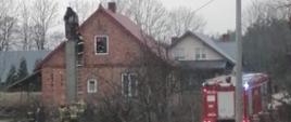 Na zdjęciu widać dom w którym doszło do pożaru w przewodzie kominowym. W oknie domu na poddaszu stoi strażak. Na drabinie stoi strażak, który wykonuje prace rozbiórkowe komina. Przy drabinie na ziemi stoi 5 strażaków, którzy zabezpieczają strażaka na drabinie. Obok domu stoi samochód pożarniczy.