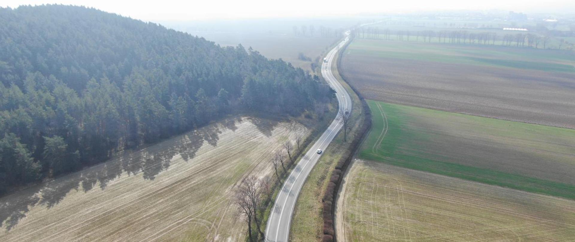 Widok z lotu ptaka na wijącą się drogę biegnącą między lasem a polami uprawnymi. Po lewej stronie znajduje się gęsty las, po prawej rozległe pola w różnych odcieniach brązu i zieleni. Na drodze jedzie pojedynczy samochód, a cała scena jest lekko spowita mgłą.