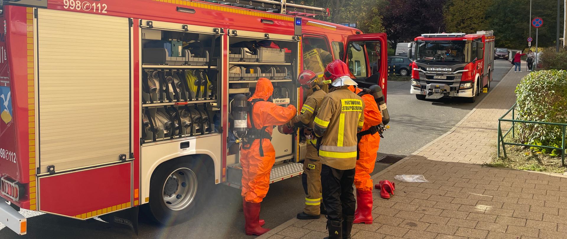 Samochody strażackie na ulicy obok bloku. Przy samochodzie strażacy ubieraja sie w specjane ubrania ochronne