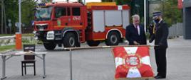 Wicewojewoda Sylwester Dąbrowski wbija pamiątkowy gwoźdź w drzewcu sztandaru. W tle widoczny duży wóz strażacki. Obok sztandaru ustawiony stolik z księgą pamiątkową.