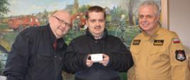 Na tle ściany, na której jest namalowany obraz przedstawiający akcję gaszenia pożaru stoją obok siebie, od lewej strony mieszkaniec powiatu rawickiego (nosi okulary), który wygrał czujnik czadu, brat zwycięzcy oraz zastępca komendanta powiatowego PSP. Mężczyźni mają na sobie kurtki w kolorze ciemnym, natomiast strażak - mundur służbowy w kolorze piaskowym. Mężczyzna stojący w środkui trzyma w rękach biały czujnik tlenku węgla. 