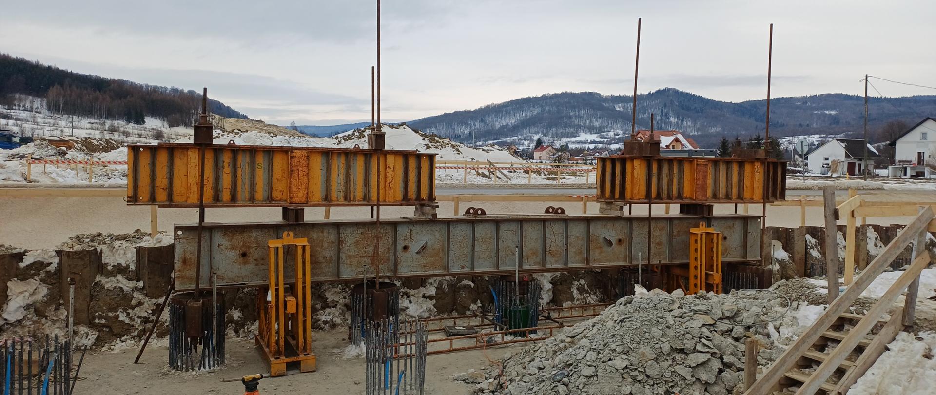 Na zdjęciu widoczne jest stanowisko przygotowane do próbnego obciążenia pali fundamentowych. W górnej części fotografii umieszczone są masywne, stalowe belki o dużym przekroju, ustawione na podporach. W dolnej części kadru widoczne są przygotowane głowice pali — wystające pionowo pręty zbrojeniowe tworzące kosze zbrojeniowe. To na nich mierzona będzie osiadanie pala pod obciążeniem.