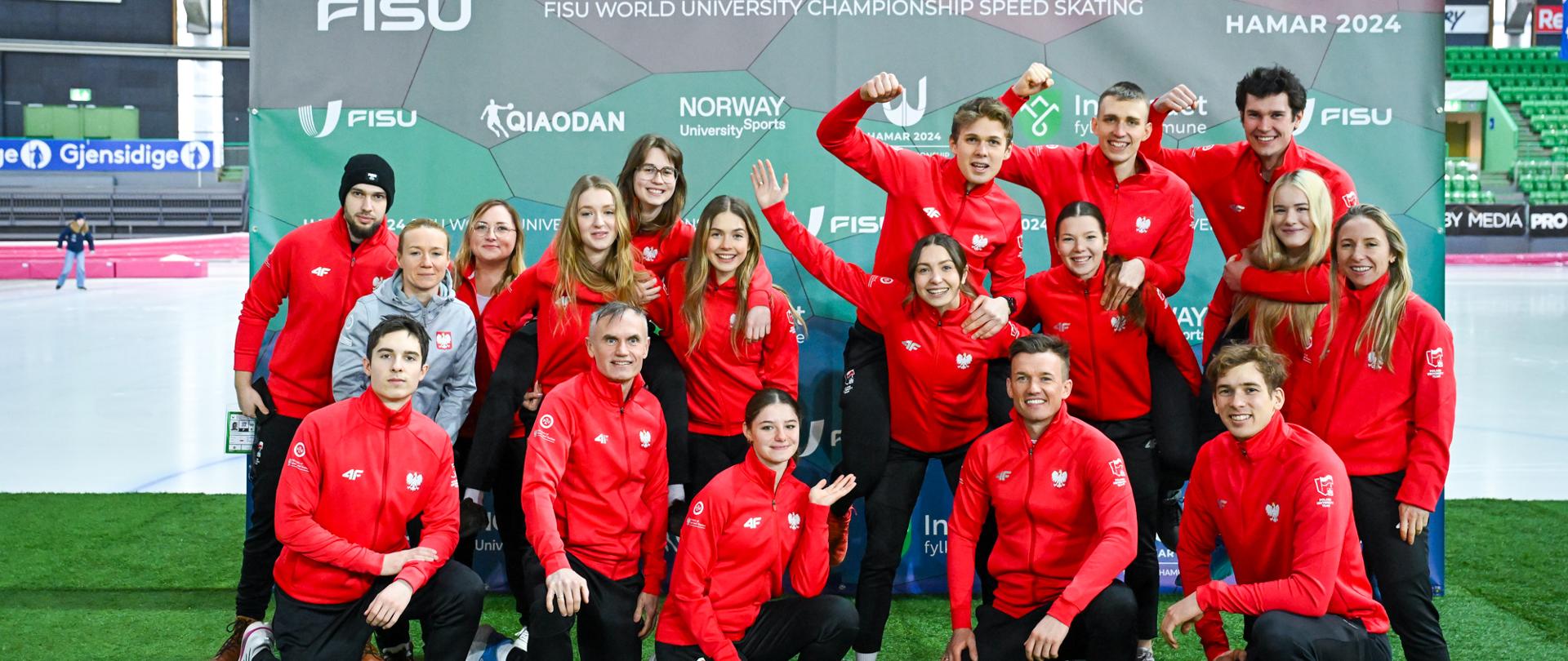 Pod ścianką z napisem Hamar 2024 stoi duża grupa osób w czarno-czerwonych strojach.