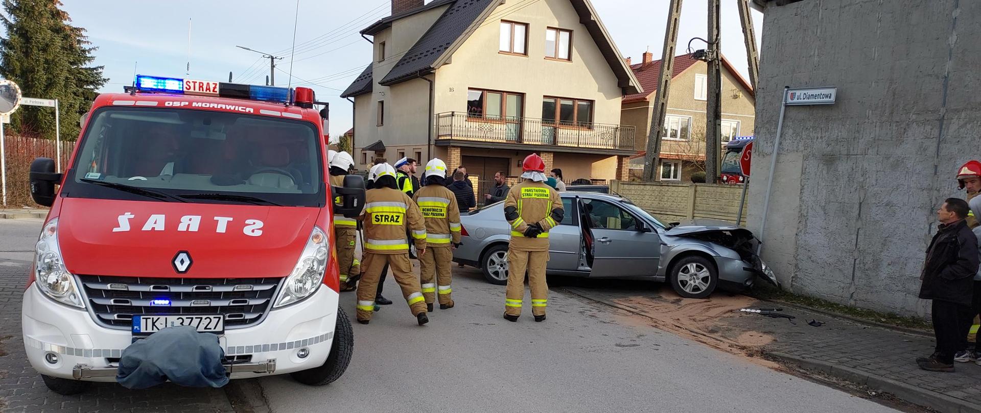 Na zdjęciu po lewej stronie widać samochód pożarniczy przy którym stoją strażacy. Z prawej stoi zniszczony samochód osobowy po zderzeniu oparty przodem o ścianę budynku. Pod autem widać rozległą plamę sorbentu do usuwania plam.
