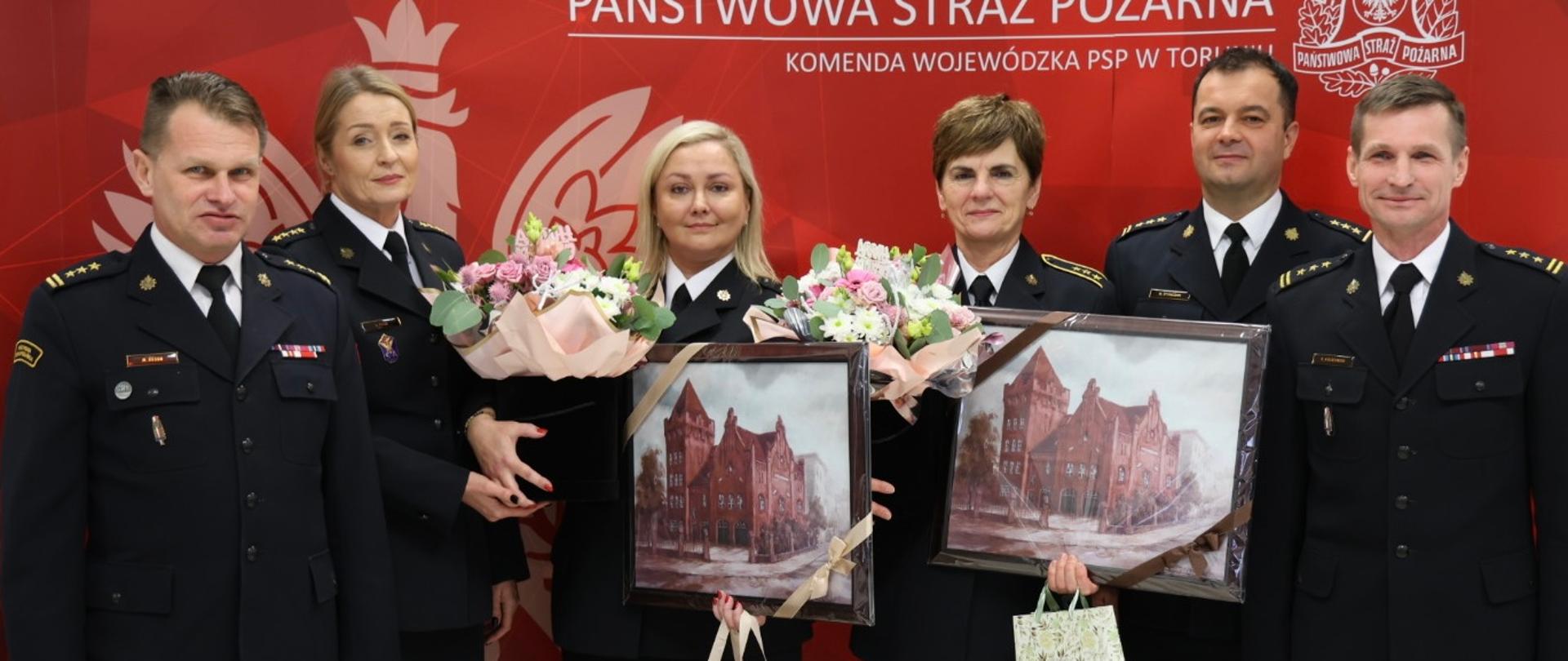 Sześcioro funkcjonariuszy w mundurach wyjściowych stoi w szeregu, pośrodku dwie funkcjonariuszki, trzymające w roku kwiaty i obrazy, a także torebki prezentowe. W tle ścianka reklamowa w kolorze czerwonym.