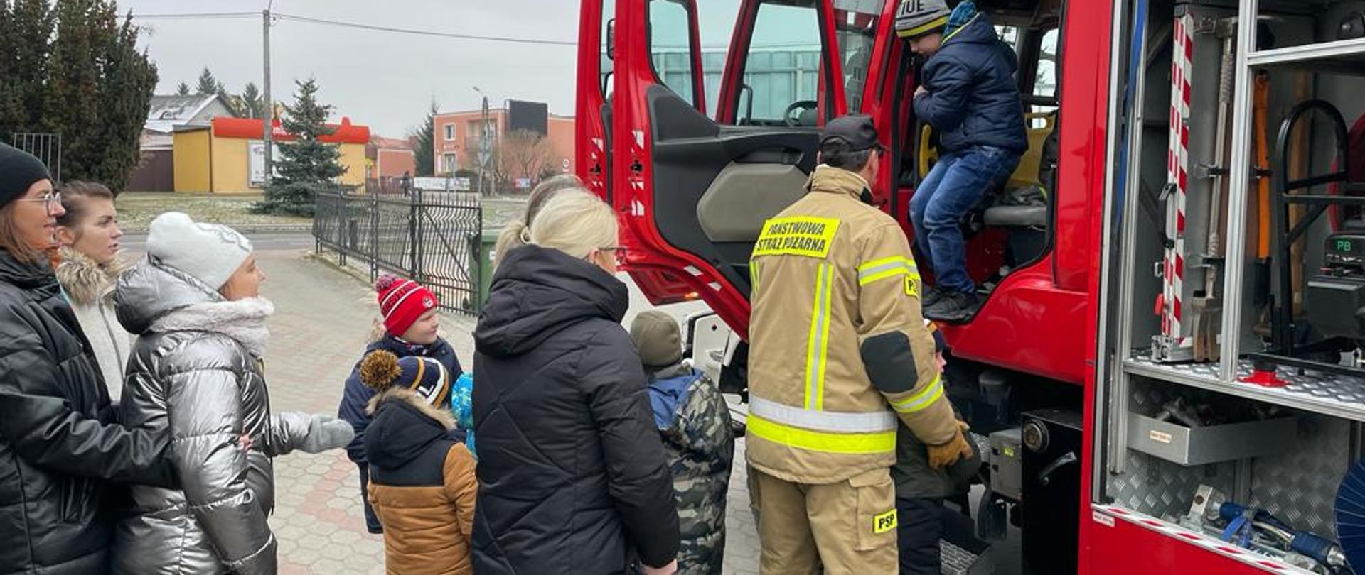 Dzieci stoją obok czerwonego, ciężarowego samochodu pożarniczego. Drzwi do samochodu sa otwarte. Jednemu z dzieci strażak pomaga wyjść z kabiny.