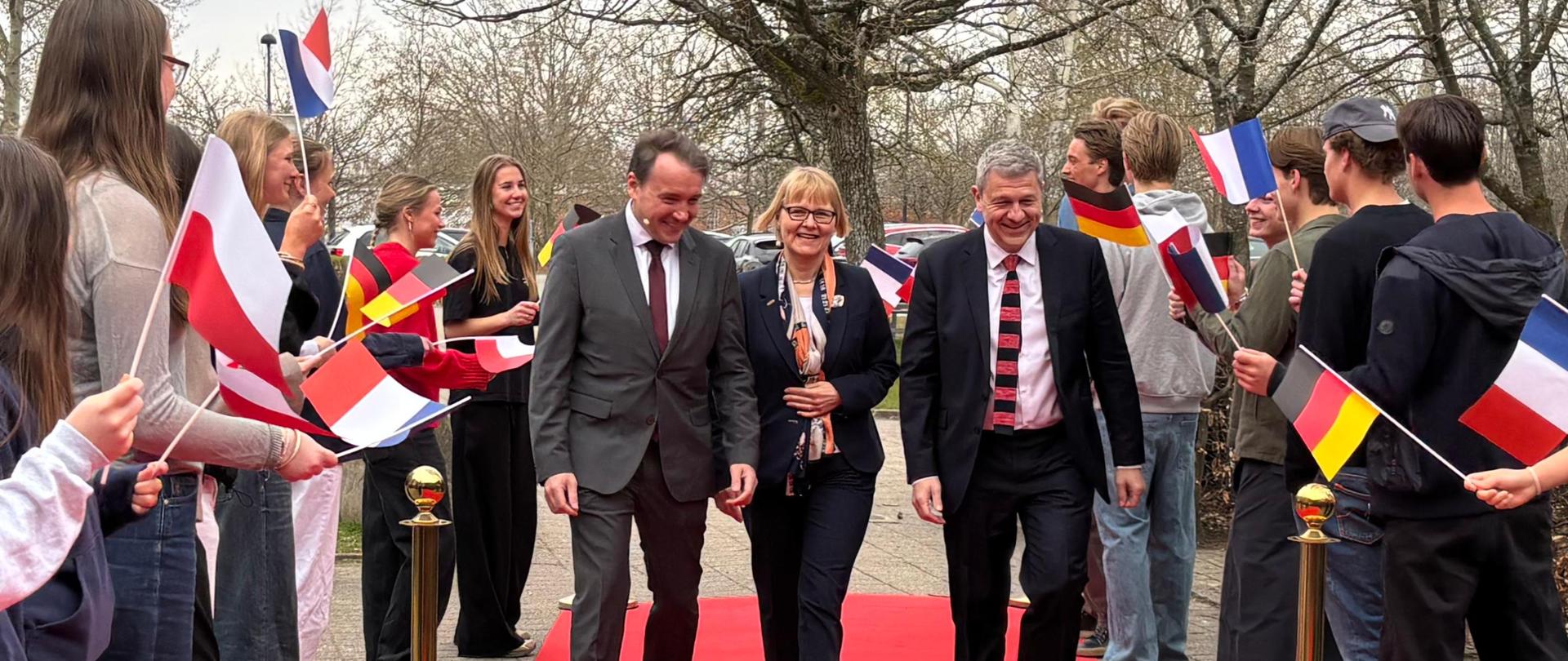 A group of people walks along a red carpet while students standing on both sides hold national flags during an official event.