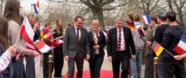 A group of people walks along a red carpet while students standing on both sides hold national flags during an official event.
