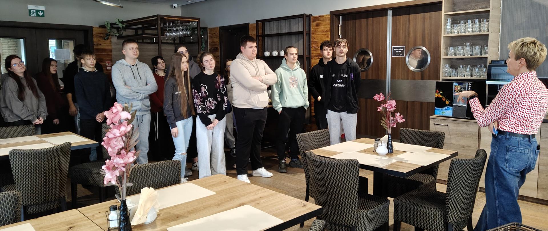 We wnętrzu nowoczesnej restauracji młodzież stoi w grupie i słucha przemówienia kobiety o jasnych, krótkich włosach.