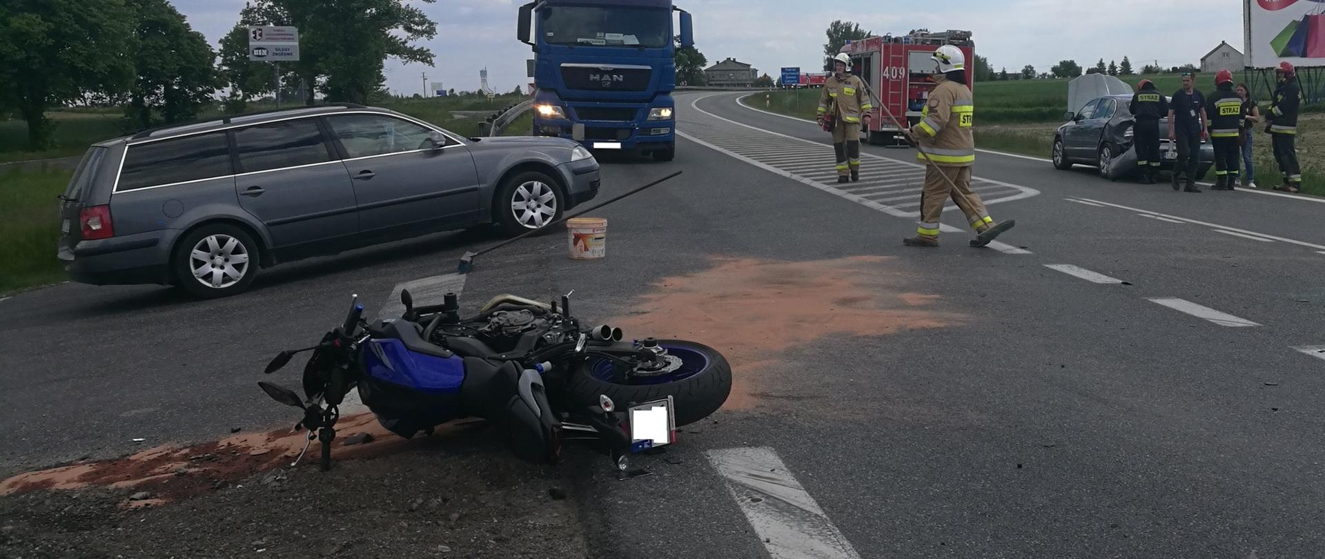 Na zdjęciu widać sytuację po zdarzeniu na DK91. Na skrzyżowaniu leży motocykl, wokół którego powstało rozlewisko płynów eksploatacyjnych. Rozlewisko pokryte sorbentem. Po drugiej stronie jezdni widać auto osobowe, które zderzyło się z motocyklem W tle widać pojazd OSP oraz ratowników prowadzących działania ratownicze.