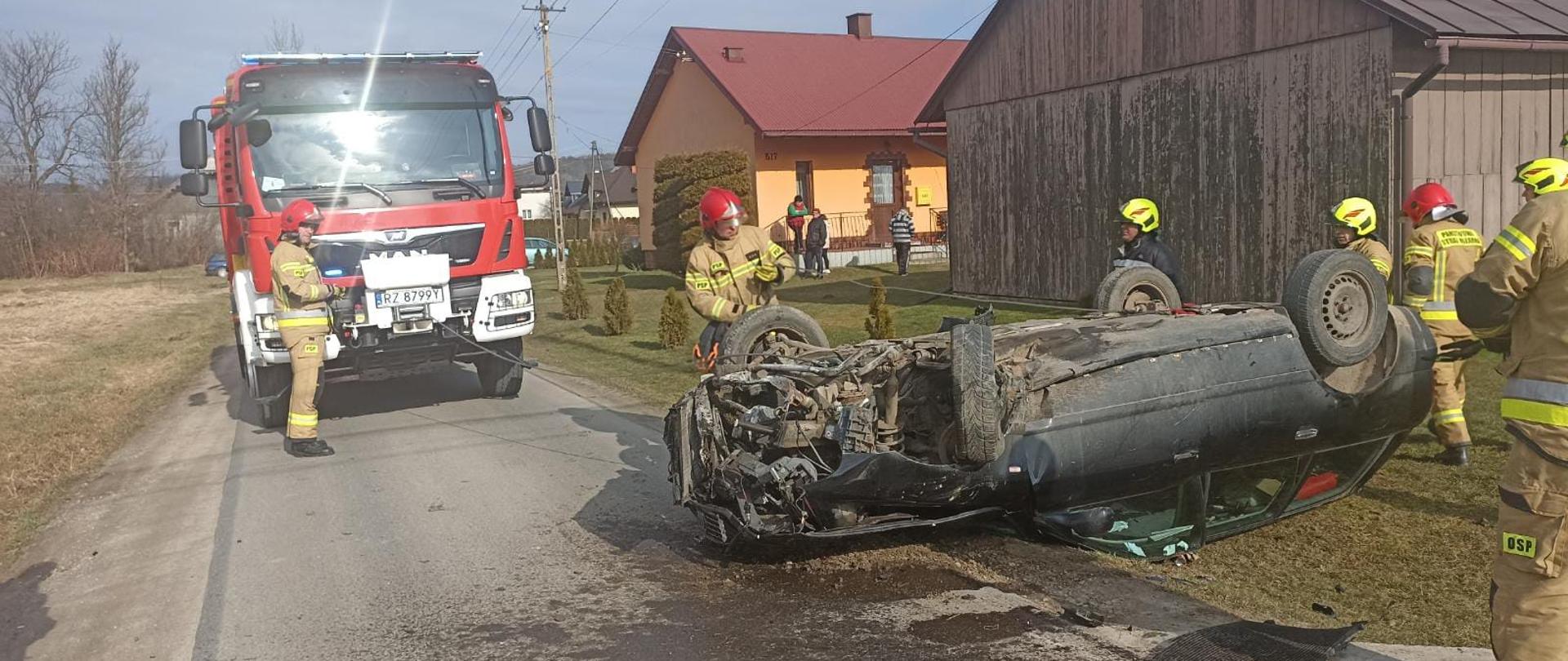 Zdjęcie przedstawia pojazd biorący udział w zdarzeniu, strażaków i inne służby oraz ich pojazdy.