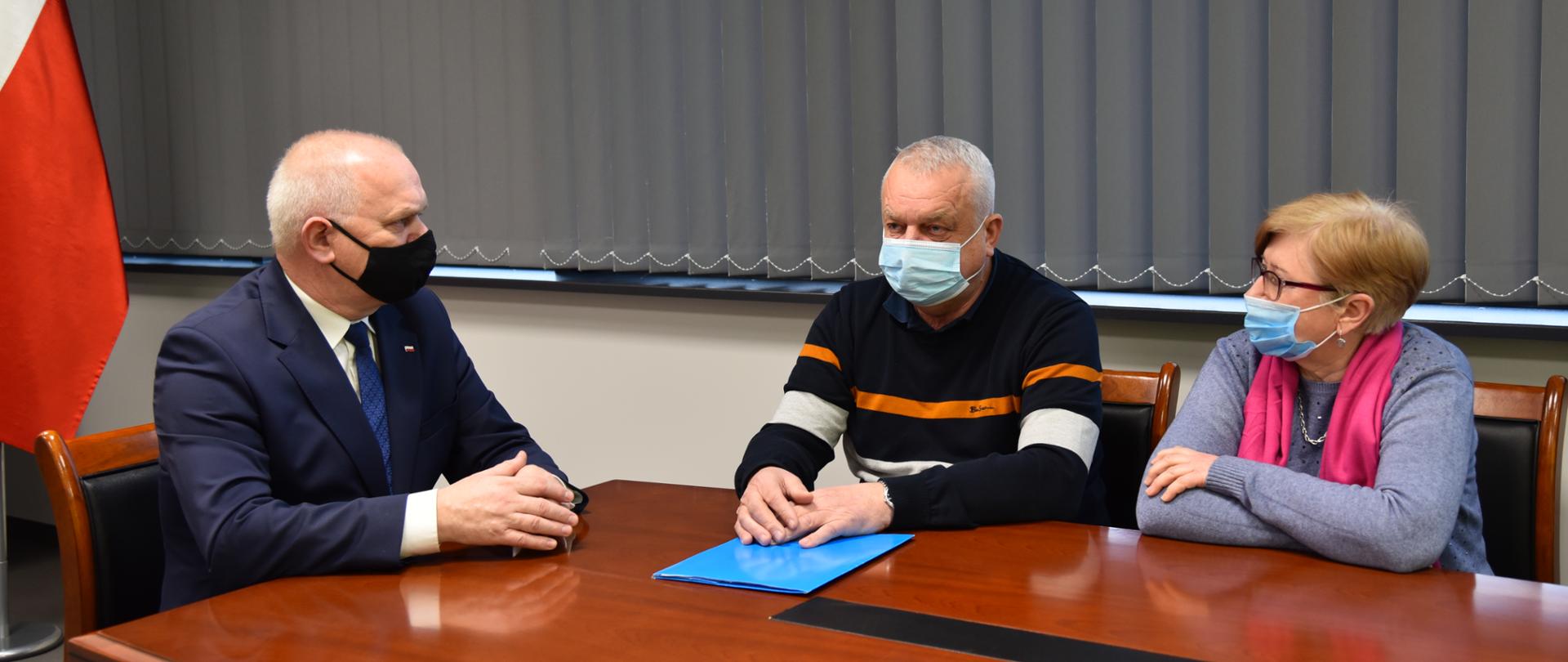 Wojewoda siedzi przy stole w gabinecie. Po jego lewej stronie, obok siebie siedzi radny i pani sołtys. W tle szare, zasłonięte rolety i skrawek biało-czerwonej flagi.