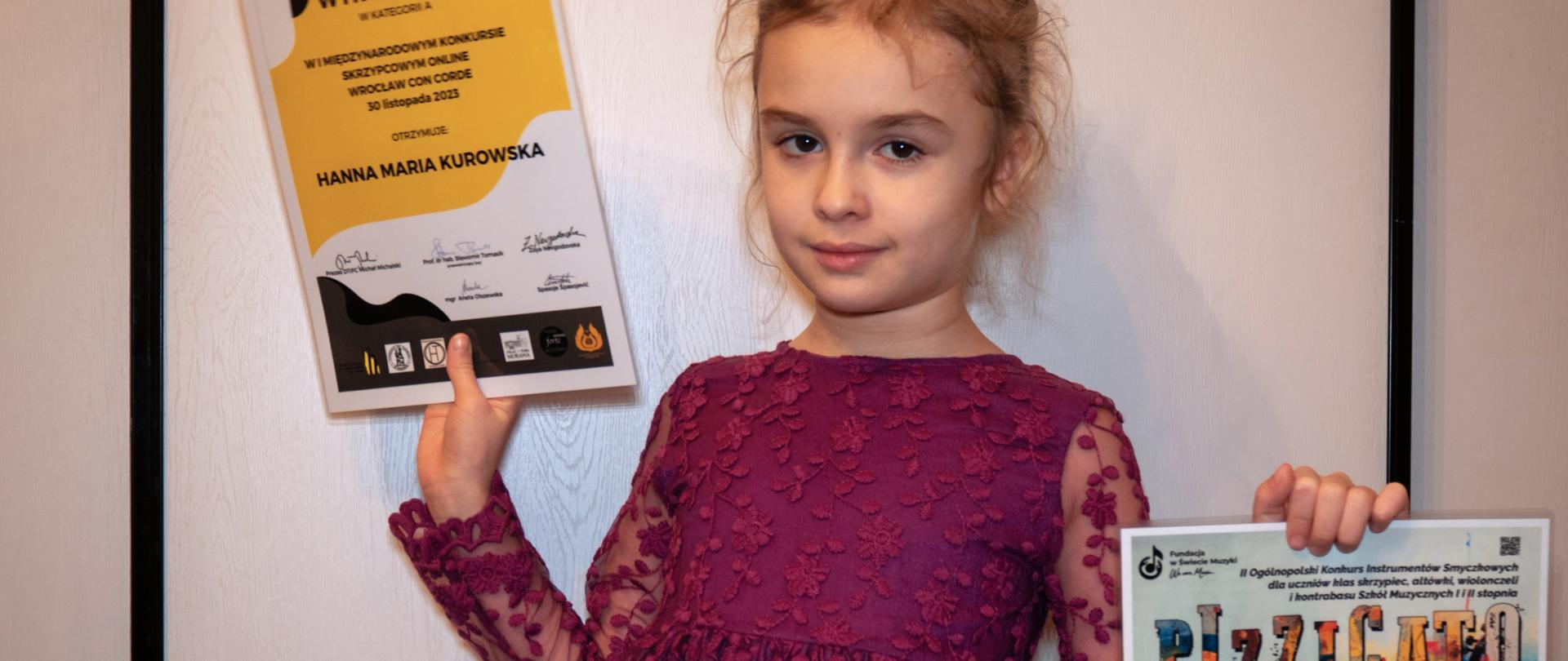 Dziewczynka stojąc trzyma w rękach dwa dyplomy.