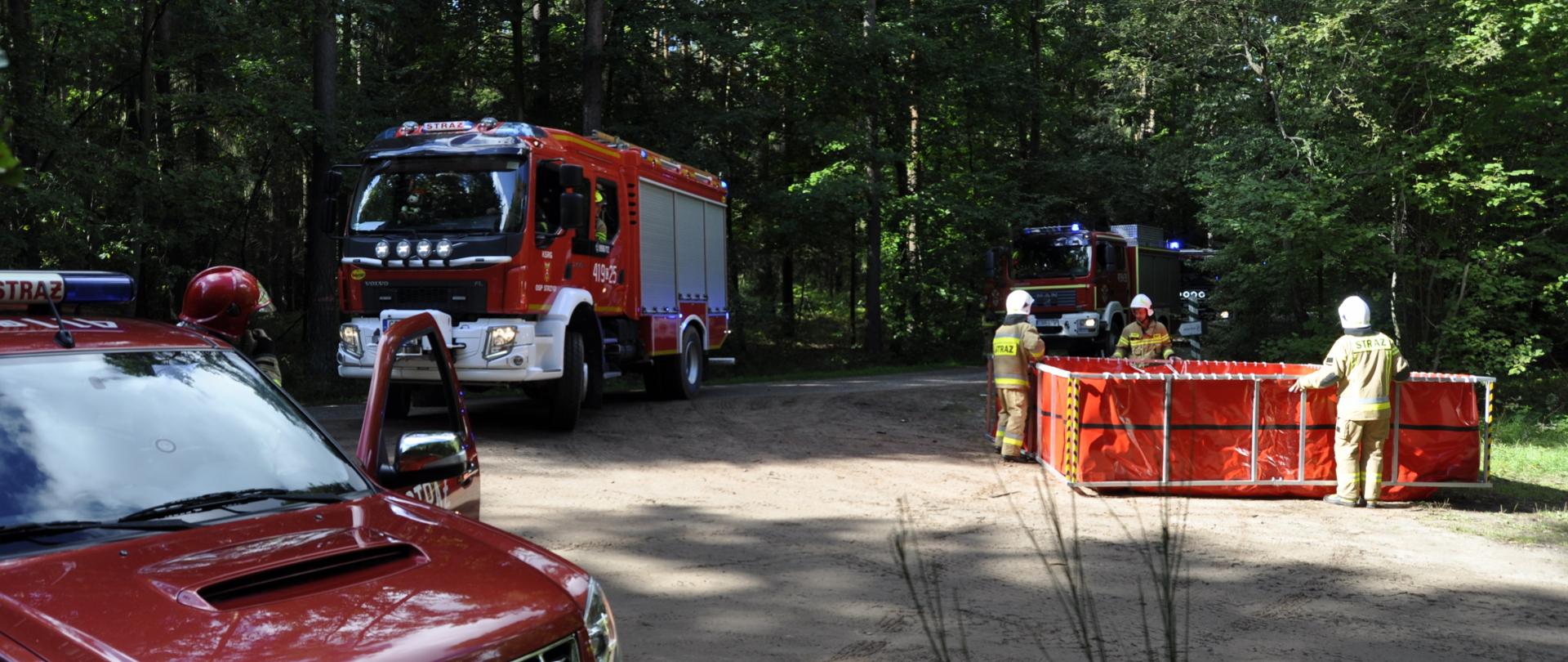 Zdjęcie przedstawia pojazdy pożarnicze dojeżdżające do zbiornika wody rozstawianego przez strażaków na leśnym skrzyżowaniu dróg.