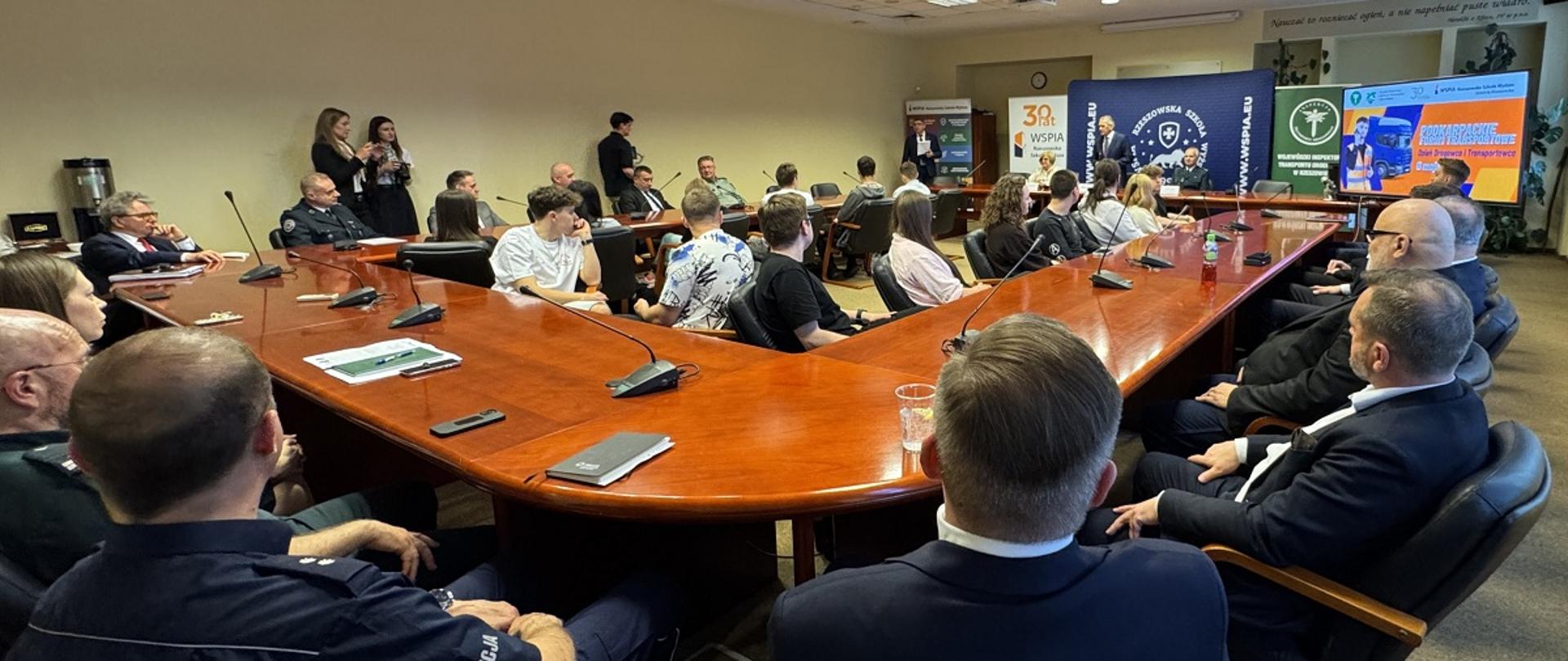 Podkarpackie Forum Transportowe. Uczestnicy siedzą w sali przy stołach. W tle przemawia Rektor WSPiA. 