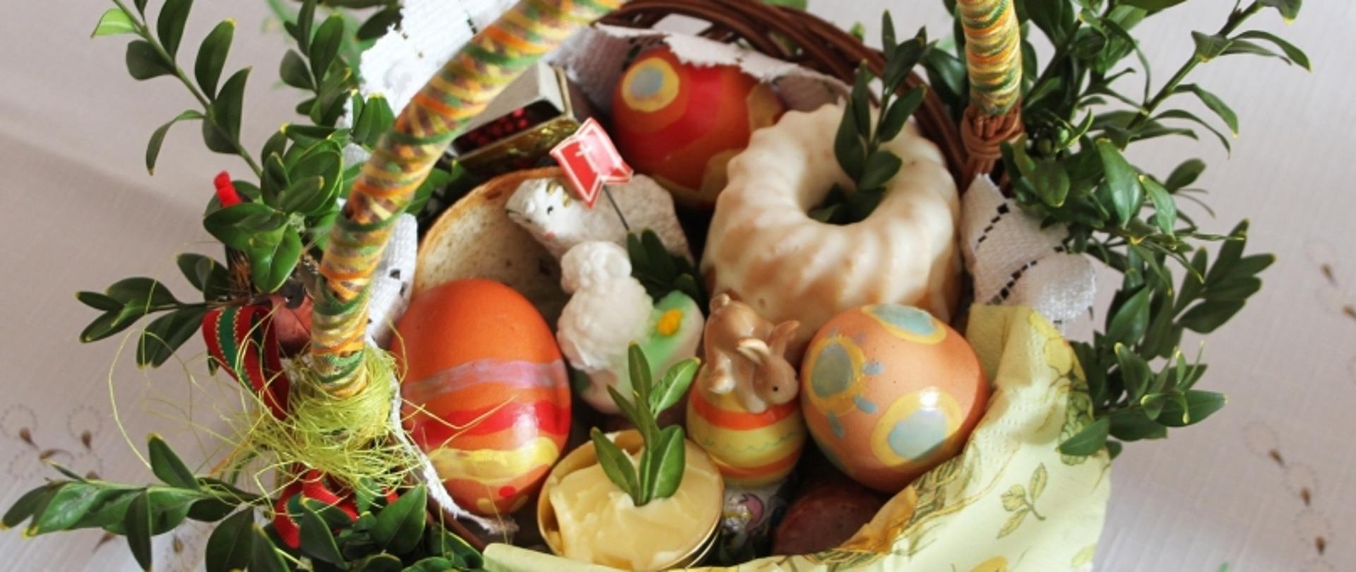 Koszyk wielkanocny z pisankami, barankiem z ciasta, ceramicznym zajączkiem i gałązkami zieleni, ozdobiony kolorową wstążką.
