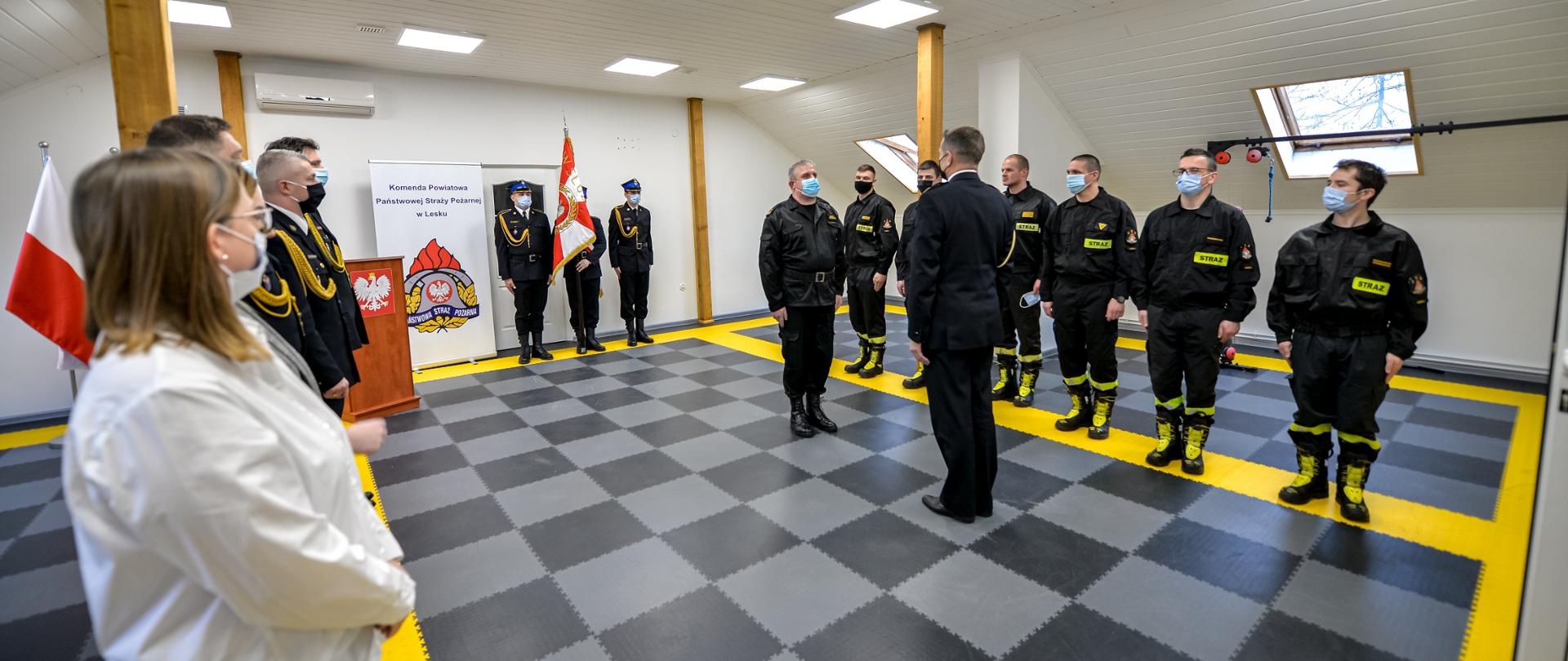 Zdjęcie przedstawia strażaków w mundurach wyjściowych i pracowników cywilnych Komendy Powiatowej Państwowej Straży Pożarnej w Lesku podczas uroczystej zbiórki z okazji przejścia na zaopatrzenie emerytalne Dowódcy Jednostki Ratowniczo – Gaśniczej w Lesku. Na zdjęciu jeden ze strażaków – dowódca uroczystości, składa meldunek Komendantowi Powiatowemu Państwowej Straży Pożarnej w Lesku. W centralnej części kadru stoi trzech strażaków w mundurach wyjściowych, jeden z nich trzyma sztandar Komendy Powiatowej Państwowej Straży Pożarnej w Lesku. Z lewej strony zdjęcia w jednym szeregu stoją strażacy w mundurach wyjściowych oraz dwie kobiety w ubraniu cywilnym. Z prawej strony również w jednym szeregu stoją strażacy w mundurach koszarowych.