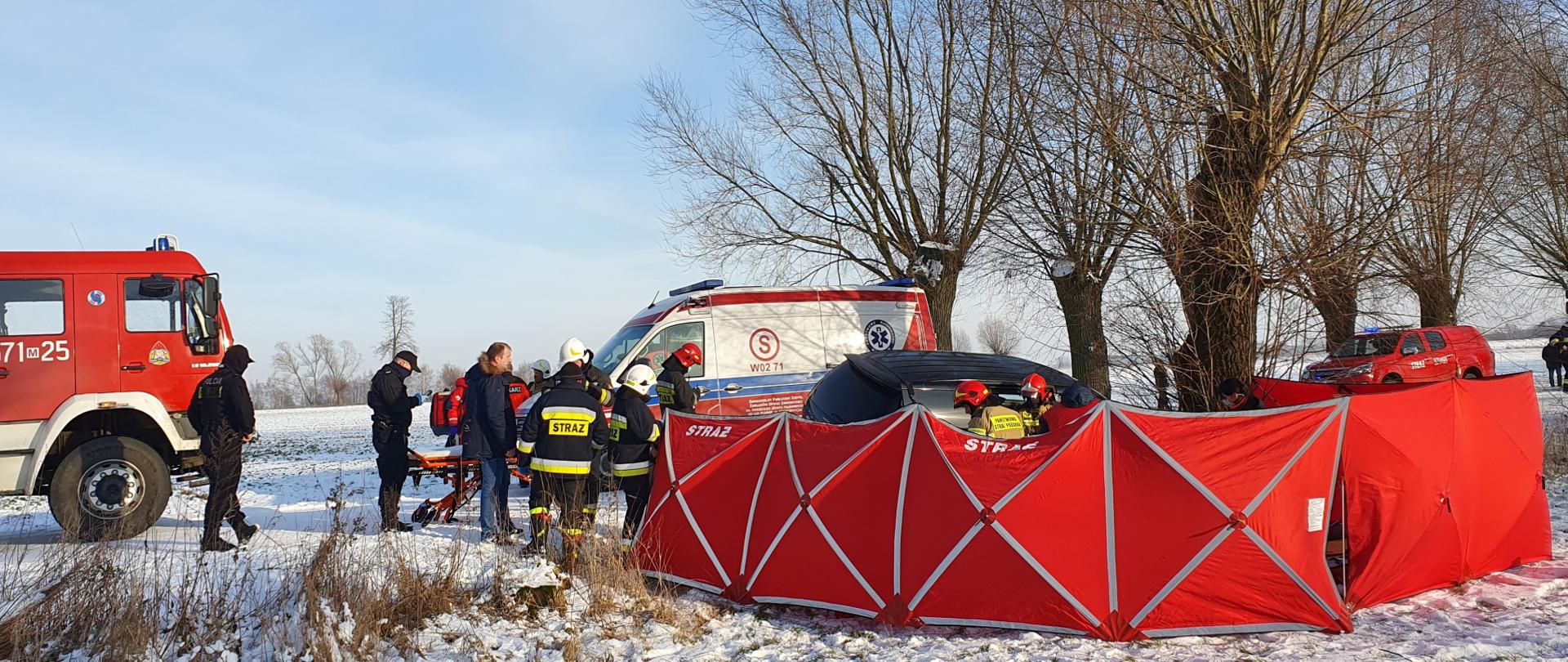 Parawan w kolorze czerwonym zasłaniający miejsce wypadku, obok stoi samochód pożarniczy i pogotowie ratunkowe a także strażacy i osoby postronne, wokół leży śnieg.
