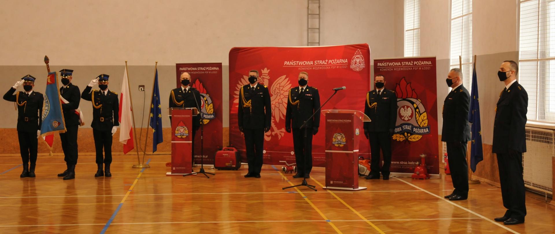 Na zdjęciu znajduje się 9 funkcjonariuszy PSP w trakcie uroczystego apelu. 3 z nich to poczet sztandarowy ze sztandarem KW PSP w Łodzi pozostali to kadra kierownicza oraz funkcjonariusze prowadzący apel, wśród nich jest nadbryg. Krzysztof Hejduk zastępca komendanta głównego PSP oraz st. bryg. Grzegorz Janowski łódzki komendant wojewódzki PSP