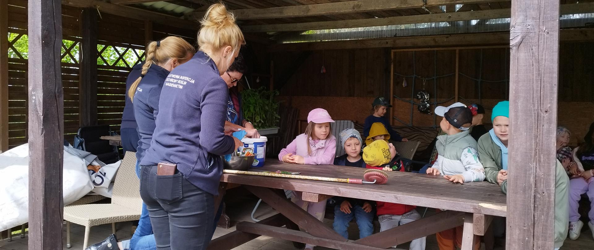 grupa dzieci siedzi pod wiata i słuchają opiekunek