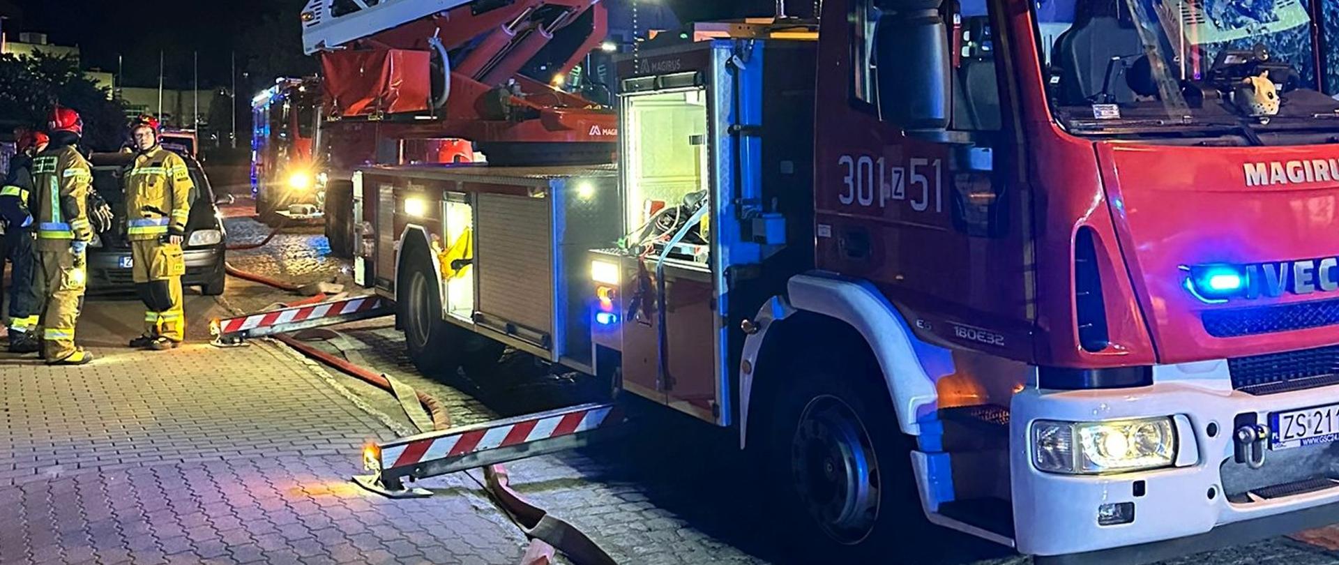 Strażacy z Komendy Miejskiej PSP w Szczecinie przy wozie z drabiną mechaniczną podczas nocnej akcji gaśniczej. Na zdjęciu widoczny pojazd pożarniczy z wysuniętymi podporami oraz ratownicy w pełnym umundurowaniu bojowym.