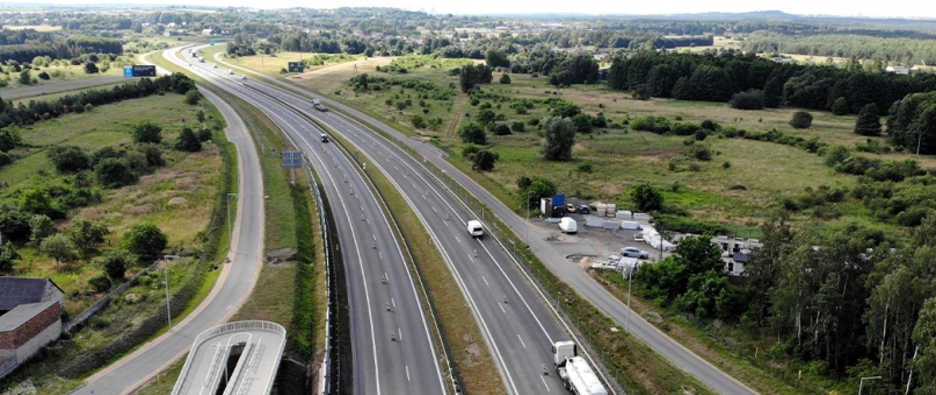 Autostrada z pojazdami w otoczeniu zieleni, z boczną drogą i zabudowaniami, widok z lotu ptaka.