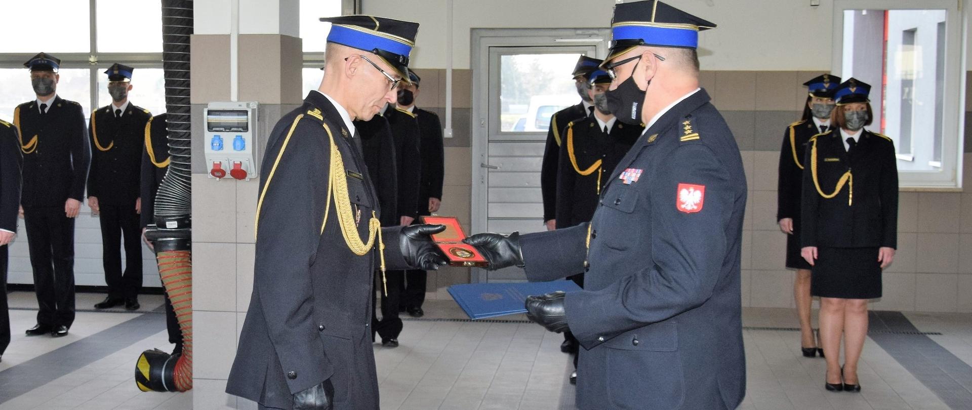Na zdjęciu stoi komendant powiatowy PSP w Wąbrzeźnie, który odbiera od komendanta wojewódzkiego PSP pamiątkowy medal. W tle pracownicy KP PSP w Wąbrzeźnie. 