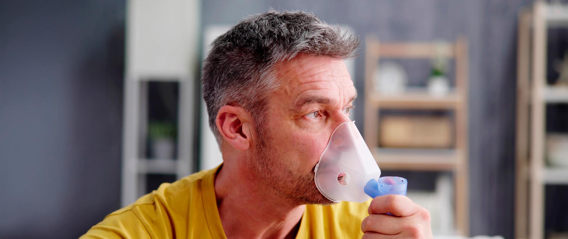 Mężczyzna z lelkko siwiejącymi włosami w żółtej koszulce przykłada do twarzy maskę nebulizatora.