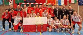 Grupa sportowców pozuje przed bramką piłkarską do wspólnego zdjęcia, w pierwszym rzędzie dwóch strażaków trzyma flagę Polski oraz stoją dwa puchary. Na ścianie za zawodnikami wiszą flagi różnych krajów