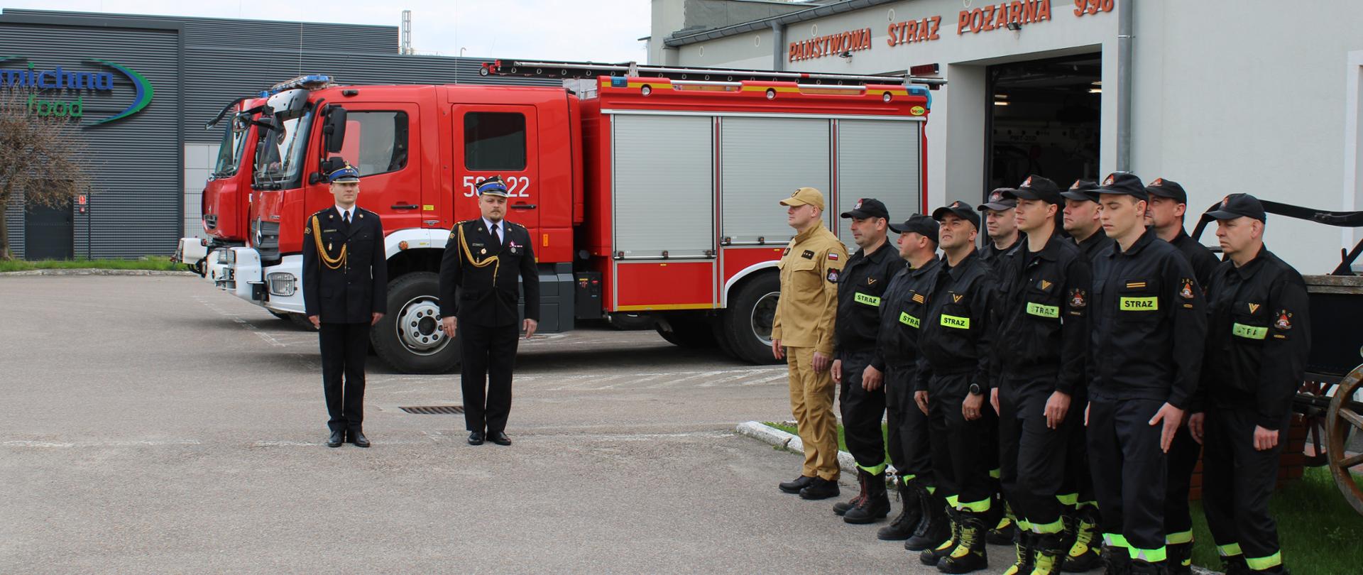 Załoga KP PSP w Nidzicy w pododdziale podczas Święta Flagi RP. Dowódca uroczystości oraz Zastępca Komendanta Powiatowego na tle pojazdów pożarniczych
