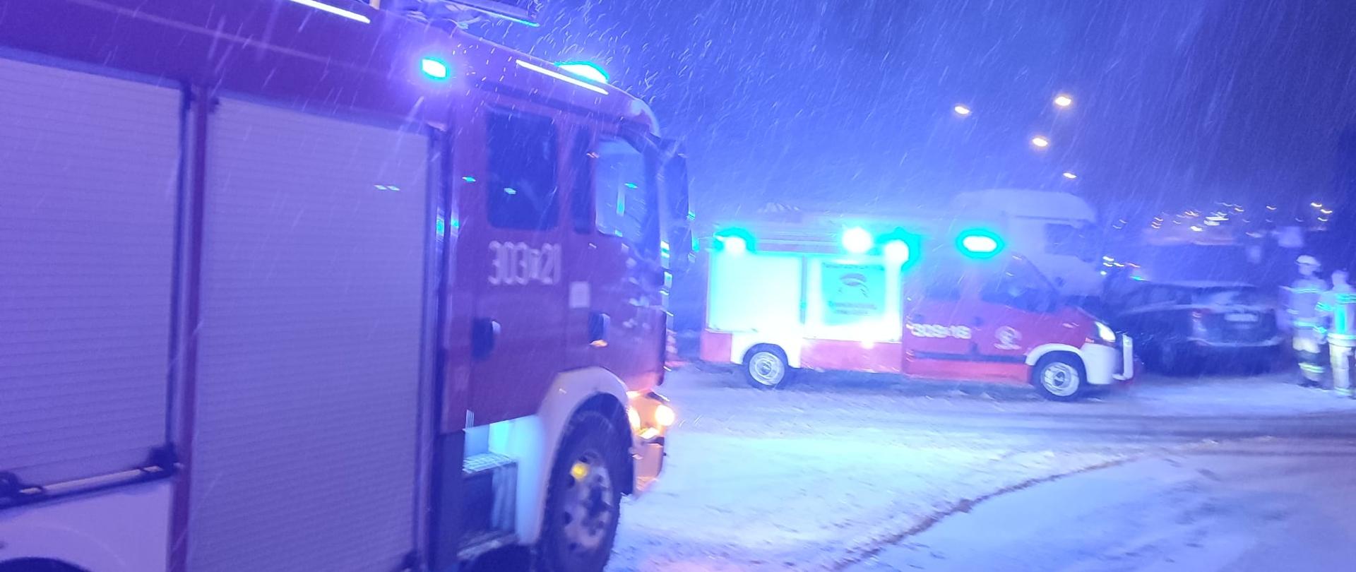 Zdjęcie przedstawia z lewej strony samochód pożarniczy i w centralnym punkcie drugi. Za nim widać fragmenty dwóch pojazdów, które uległy zderzeniu. Przy nich stoi trzech strażaków. Widać padający gęsty śnieg.