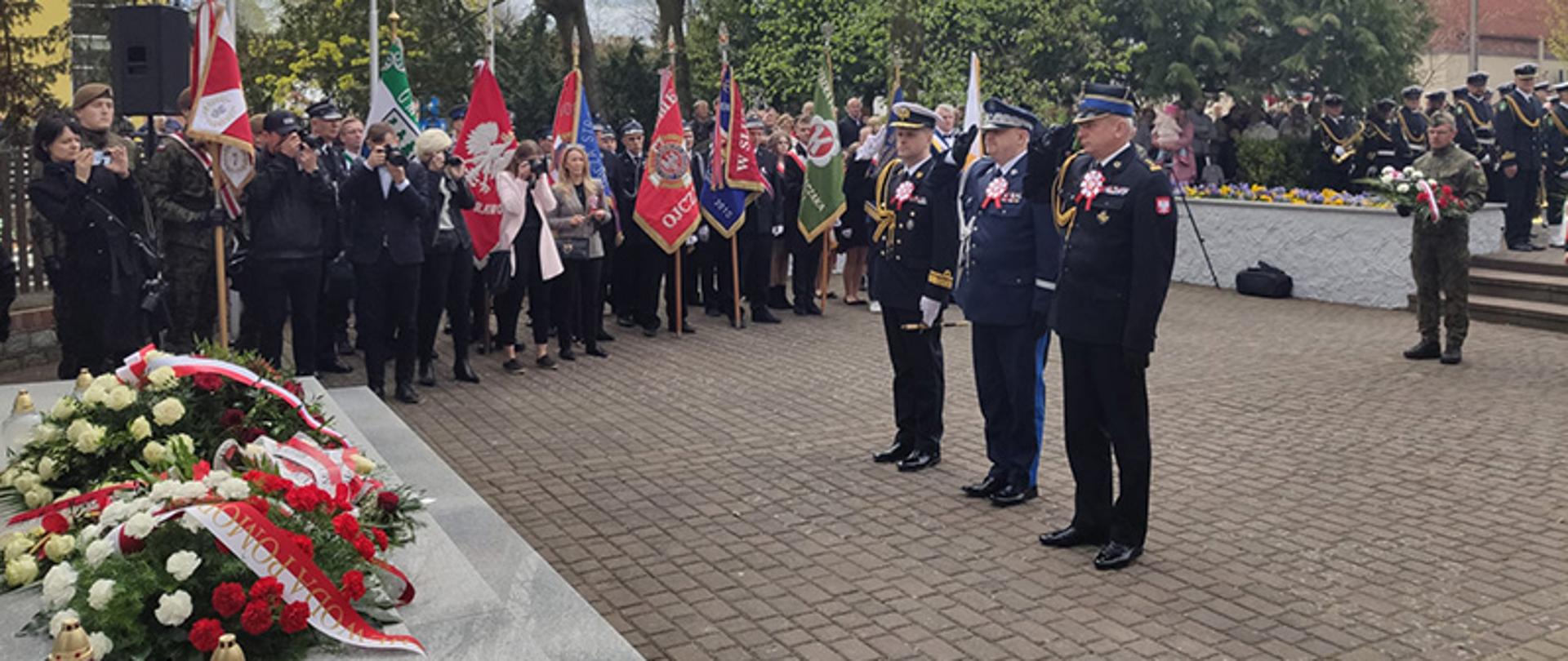 Trzech przedstawicieli służb mundurowych oddaje honor, stojąc przed pomnikiem, obok stoją poczty sztandarowe.