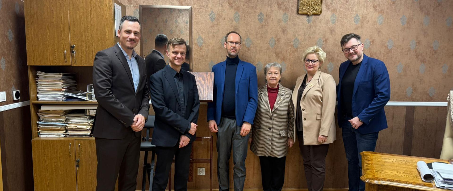 Grupa siedmiu dorosłych osób stojących obok siebie w pomieszczeniu o oficjalnym charakterze. Osoby ubrane są elegancko, w marynarki i garnitury. W tle widoczne są drewniane meble, szafka z dokumentami oraz godło Polski na ścianie. Wszyscy pozują do wspólnego zdjęcia, patrząc w stronę aparatu.