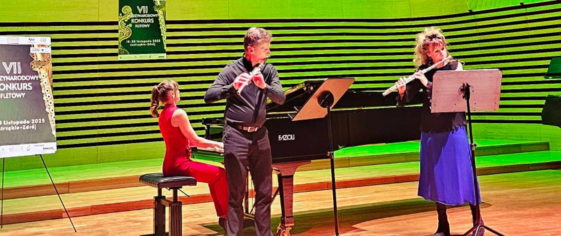 Kobieta i mężczyzna grają na fletach, kobieta akompaniuje im na fortepianie.