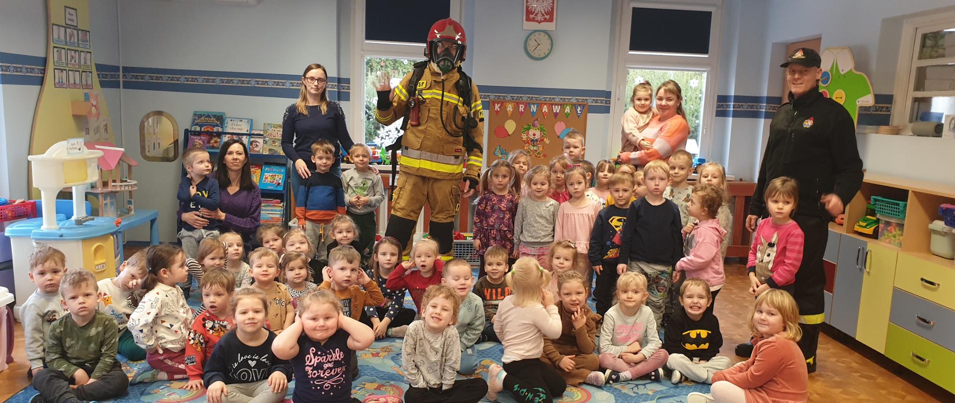 Widać dzieci z przedszkola, siedzą na dywanie, z tyłu strażak w mundurze i trzy wychowawczynie z dzieckiem na ręku, po prawej strażak