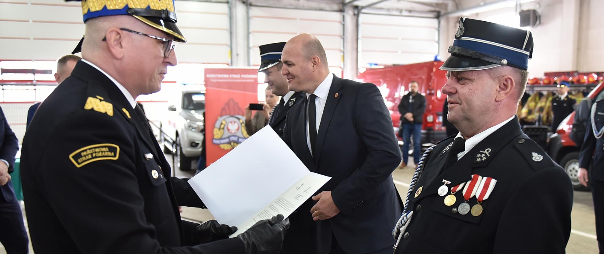 na głównym planie generał straży pożarnej wręcza decyzję strażakowi ochotnikowi, w tle inne osoby, zdjęcie wykonane w garażu