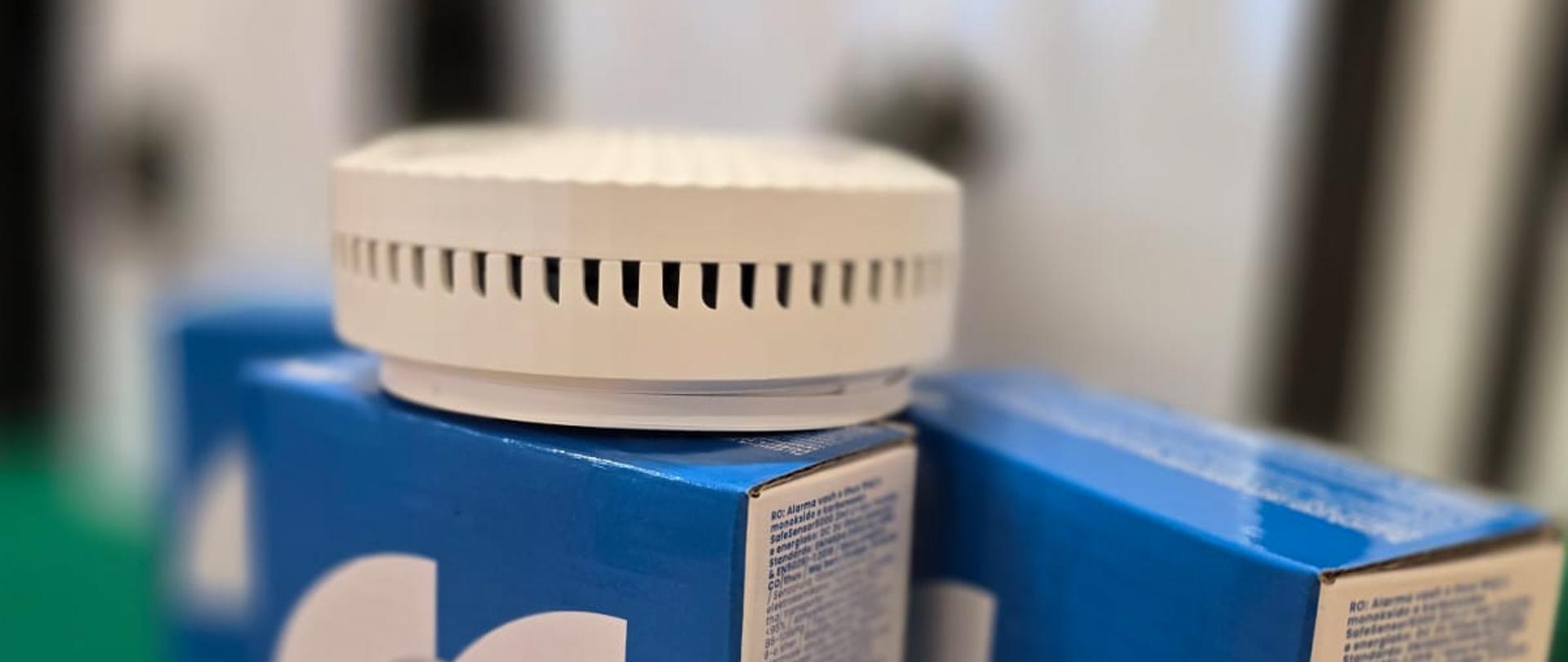 Na zdjęciu widać czujkę czadu i dymu na stoliku z zielonym obrusem.