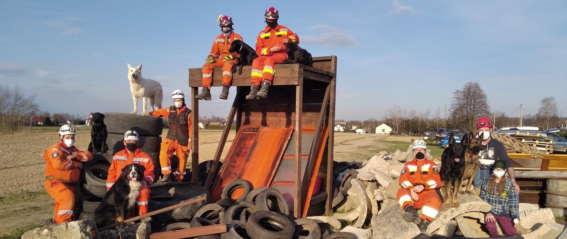 Poligon gruzowiskowy. Na pierwszym planie betonowe gruzy i sterta opon. W głębi na stertach opon i drewnianym wysokim podeście kilka zespołów ratowniczych. W zespole ratownik w pomarańczowym skafandrze i pies.