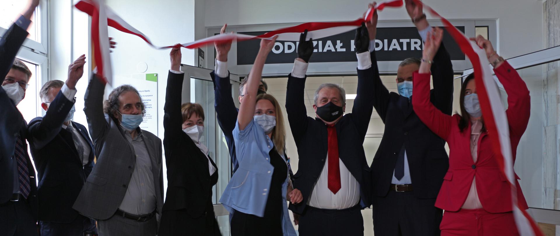 Na zdjęciu 9 osób, ustawione w rzędzie. Pozują do zdjęcia. Wszystkie osoby, ubrane elegancko, podnoszą ręce do góry, trzymają oni biało-czerwoną wstęge na znak uruchomienia oddziału pediatrii. W środkowej części kadru wojewoda opolski. W tle drzwi oddziału z napisem Oddział Pediatrii. 