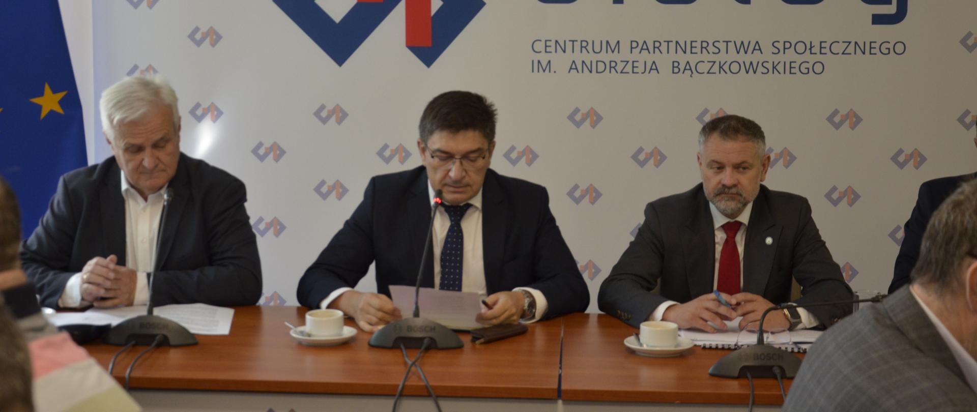 Na zdjęciu stół prezydialny za którym siedzą od lewej strony: Mariusz Kamiński – Przewodniczący Zrzeszenia Związków Zawodowych Energetyki, Wojciech Ilni8cki – Współprzewodniczący Zespołu oraz Grzegorz Wrona - Podsekretarz Stanu w Ministerstwie Aktywów Państwowych. W tle siedzących baner z napisem Centrum Partnerstwa Społecznego Dialog.