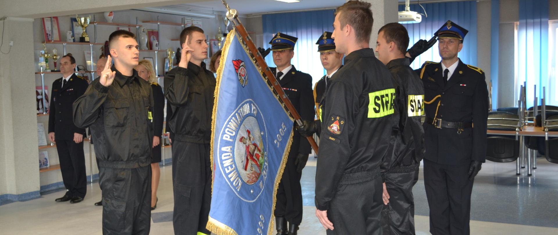 Na pierwszym planie widoczni są strażacy składający ślubowanie przy poczcie sztandarowym. Poczet składa się z trzech strażaków z czego środkowy trzyma opuszczony poczet. W tle widoczne są osoby uczestniczące w apelu