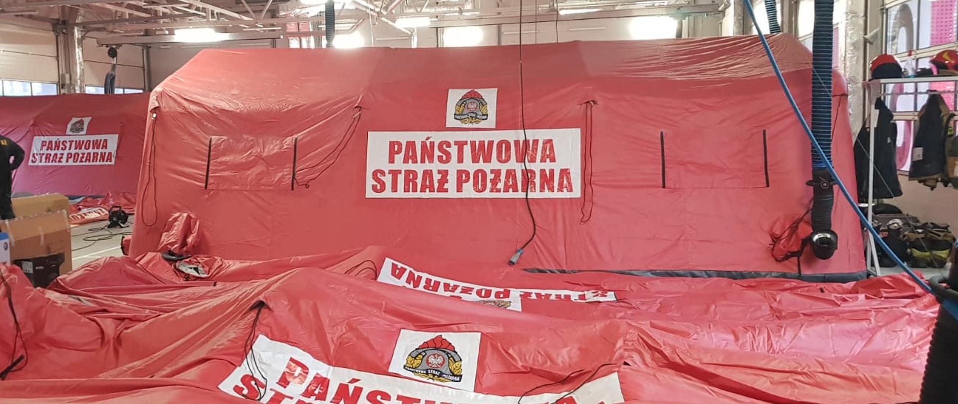 Przekazanie namiotów pneumatycznych dla mazowieckich strażaków. Na zdjęciu w garażu dwa namioty pneumatyczne pomarańczowe jeden rozstawiony z tyłu z przodu rozłożony jeszcze nie napompowany. z lewej strony stoją 3 funkcjonariusze w mundurach.