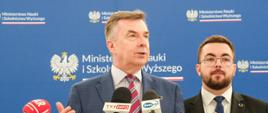 Minister Wieczorek mówi do kilku mikrofonów trzymanych przez dziennikarzy poza kadrem, obok stoi mężczyzna w garniturze, za nimi granatowa ścianka z napisem Ministerstwo Nauki i Szkolnictwa Wyższego.