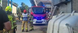 Strażacy stoją terenie gospodarstwa. W tle stoi drabina mechaniczna. 