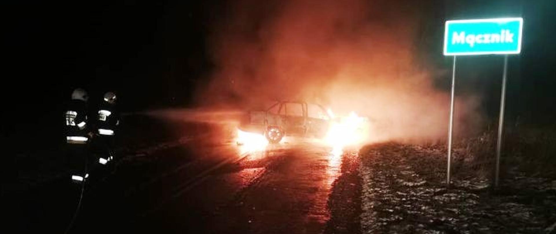 Samochód osobowy na jezdni objęty płomieniami. Dwóch strażaków trzymających wąż gaśniczy próbujących ugasić pożar pianą. Na poboczu widoczny znak drogowy z napisem Mącznik.