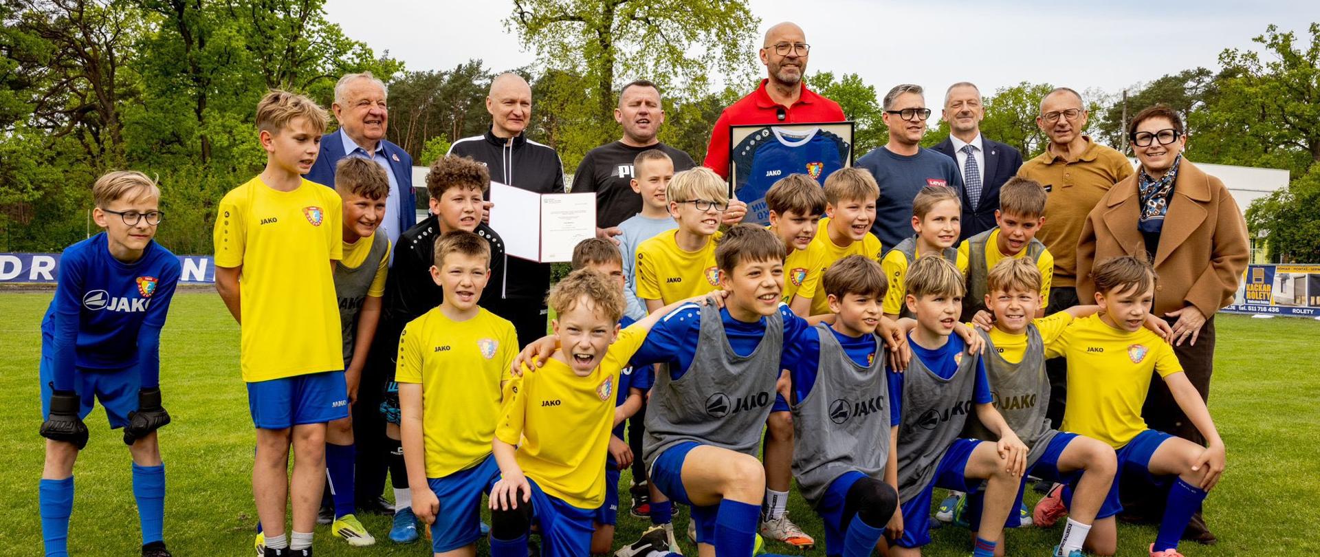 Zdjęcie zbiorowe przedstawiające ministra sportu i turystyki Jakuba Rutnickiego z młodymi piłkarzami z Drezednka. Zdjęcie zrobione na boisku piłkarskim.